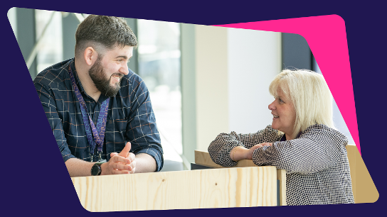Two people conversing indoors, leaning on a wooden surface with large windows in the background.