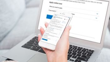 Person holding a smartphone with a website visible, laptop showing similar content in background.