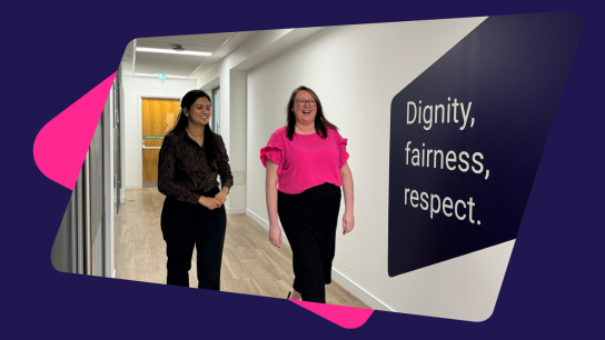 Two people walking down a bright office hallway with a wall sign that reads ‘Dignity, fairness, respect.