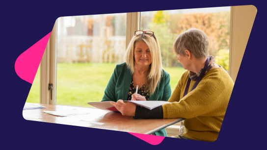 Two people sitting at a table near large windows, reviewing documents together.