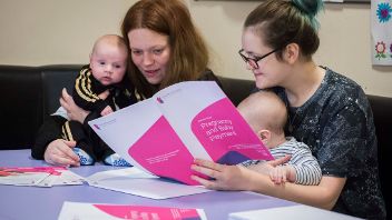 Two adults seated with babies, reviewing a booklet titled 'Pregnancy and Baby Payment'.