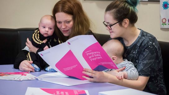 Two adults seated with babies, reviewing a booklet titled 'Pregnancy and Baby Payment'.