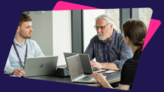 Three people seated at a table with laptops, engaged in discussion in a bright office setting.