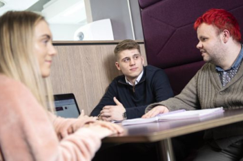 Three people seated around a table in an office, engaged in conversation with a laptop open.