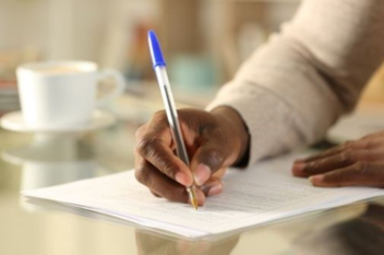Individual writing on paper with a blue pen at a table, coffee cup nearby.