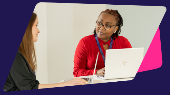 Two people sitting at a table with laptops, engaged in discussion in a bright office setting.
