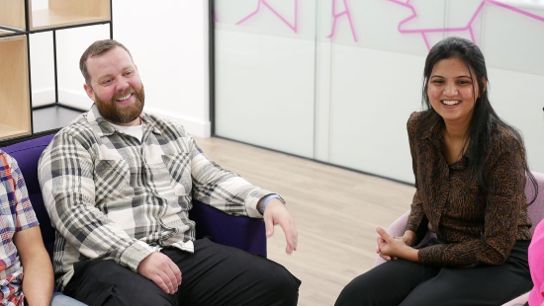 Three people seated and conversing in a modern room with glass walls and shelves.