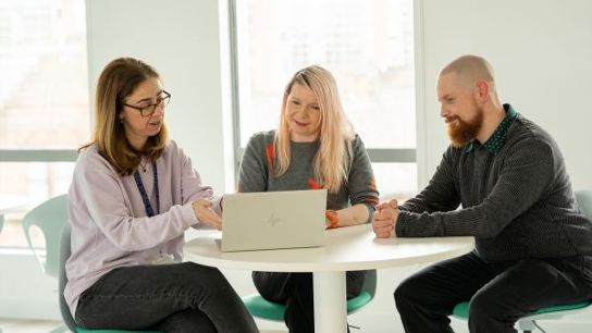 Three people seated around a table, collaborating while looking at a laptop screen.