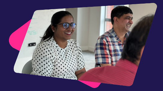 Three people seated in a meeting room, two smiling and engaged in discussion, with a whiteboard in the background.