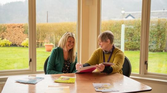 Two people seated near large windows, discussing contents of an open folder.