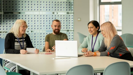 Four people around a table with laptops and documents, collaborating.
