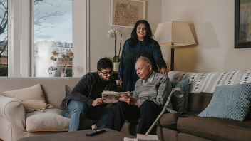 A carer is standing in a living room behind a younger relative and older relative who are sitting down.