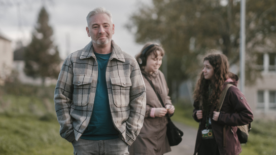 A carer is standing outside in front of the two people they care for