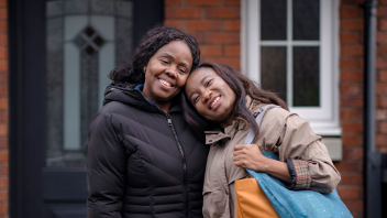 Two people standing outside a brick house, smiling and dressed warmly.