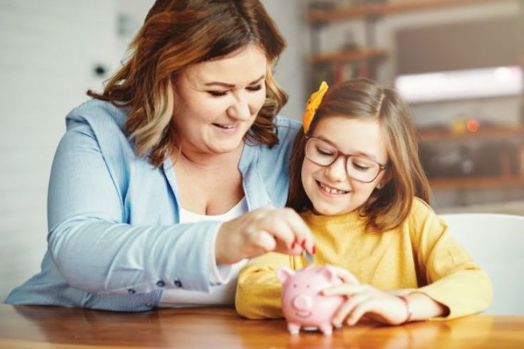 A woman and child sitting at a table. The woman is putting money into a piggy bank