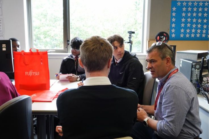 A group of school students are sat at a table listening to a visitor