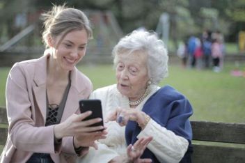 Two people seated on a bench outdoors, one showing a smartphone to the other, with trees and people in the background.