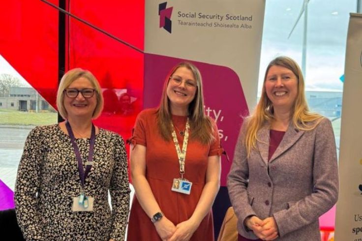 Three colleagues standing together in front of  Social Security Scotland pop up banner
