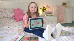 Child sitting on a bed holding a tablet displaying “Level Completed,” with toys and books nearby.
