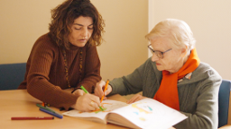 Two people sitting at a table colouring in a large book with coloured pencils.