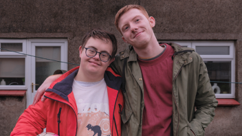 Two people standing close together outdoors in front of a building, one with an arm around the other.