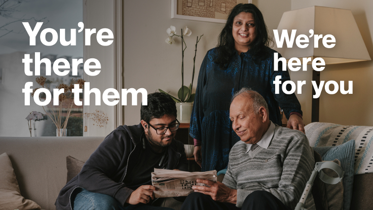 A carer is standing in a living room behind a younger relative and older relative who are sitting down. Text reads: You're there for them. We're here for you.