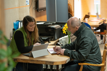 Two people sat at a table, one using a tablet and the other filling out a form
