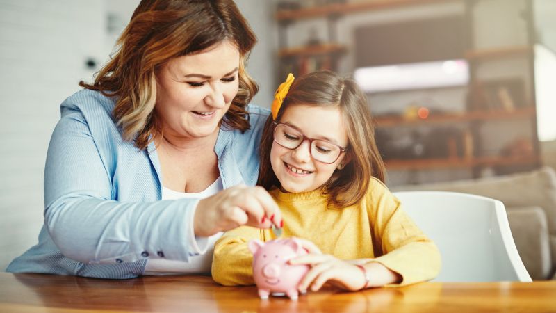 A person is with a child putting money in a piggy bank