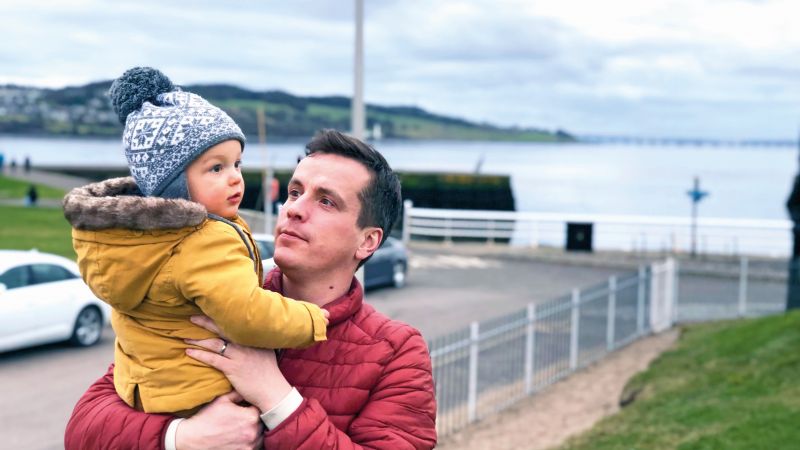 Man in a red jacket holding a baby in a yellow jacket and wooly hat