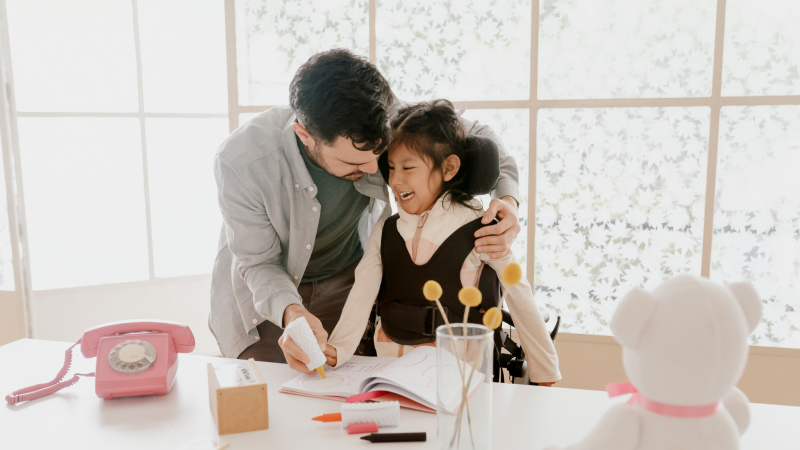 Man helping girl in wheelchair draw in colouring book