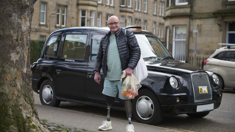 A person with prosthetic legs is standing in front of a taxi holding a shopping bag