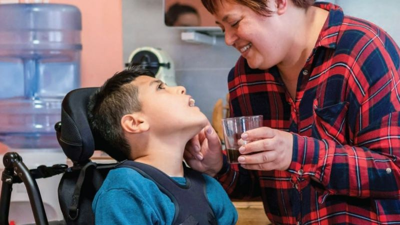 An unpaid carer helps a young boy in a wheelchair drink from a glass