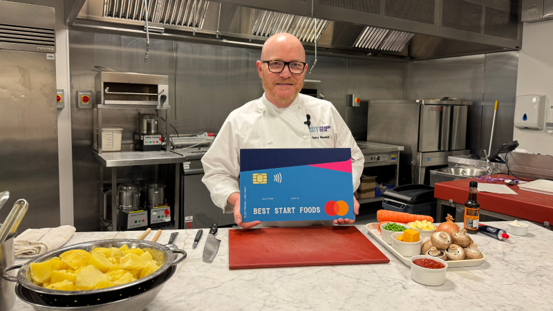 National Chef Gary Maclean is standing in a kitchen holding a giant Best Start Foods card