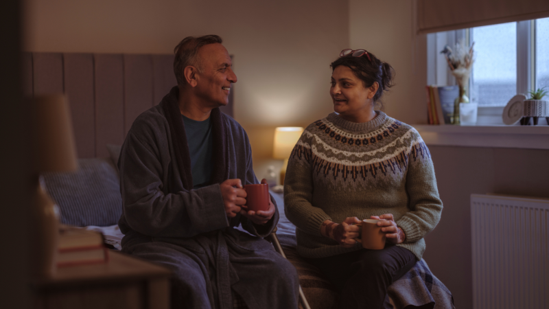 Two people are sitting on the end of a bed smiling at each other.
