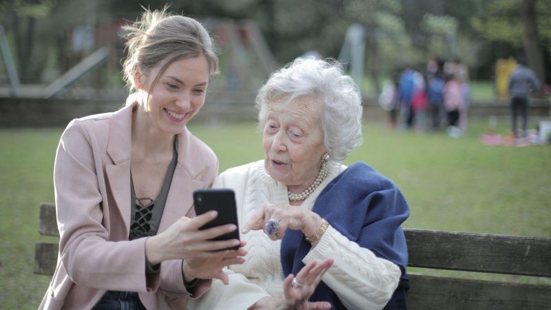 Two people, one who is elderly, sit on a park bench looking at a phone