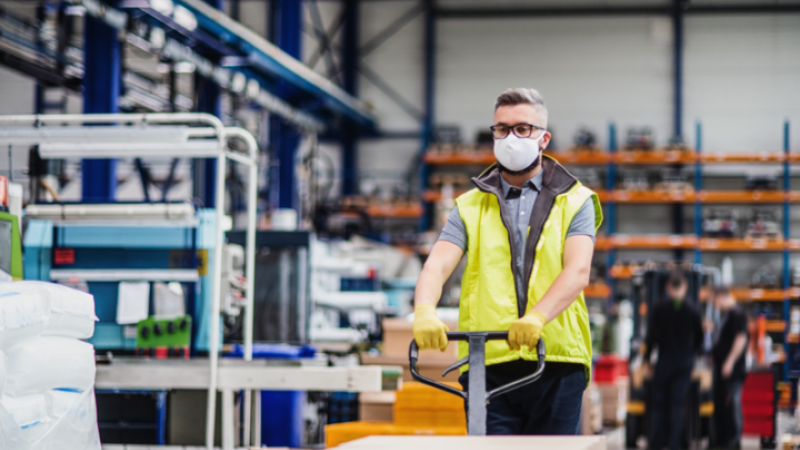 A warehouse worker is wearing a face mask and pushing a trolley with boxes on it.