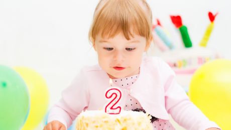 A little girl in a pink dress is standing behind a birthday cake with a number 2 candle. She is surrounded by balloons.