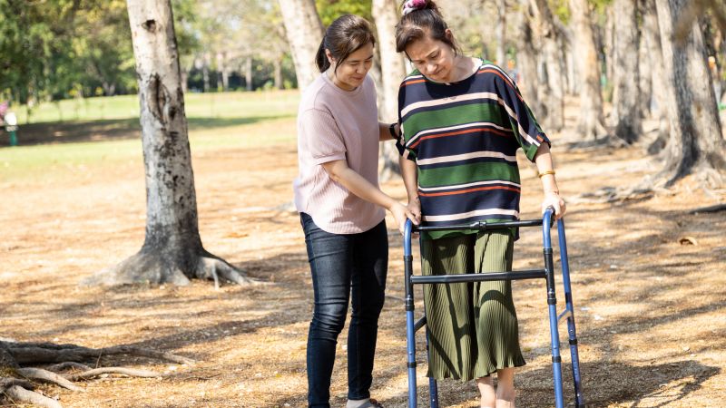 A woman is outside using a walking frame. She is being supported by a young carer.