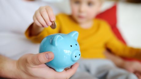 Child is sitting on his parents knee placing a coin into a piggy bank.