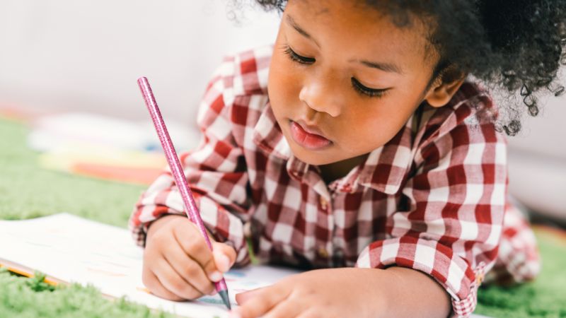 Young child lies on carpet writing in a notepad.