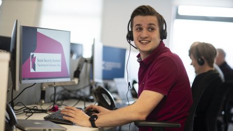 Young Social Security Scotland Client Adviser wearing headset is sitting at a desk.